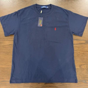 Mörkblå t-shirt - Klassisk mörkblå t-shirt med röd broderad logga på bröstet. T-shirten har rund halsringning och korta ärmar. Tillverkad i mjuk bomull för en bekväm känsla och stilren look.