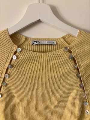 Gul ribbad stickad tröja från Zara - Trendig gul stickad tröja från Zara med ribbad struktur och dekorativa silverfärgade knappar längs axlarna. Tröjan har rund halsringning och långa ärmar. Perfekt för dig som vill ha en färgklick i garderoben. Liten i storlek så är mer som en S💛💛