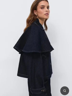 Mörkblå jeansjacka med cape - Unik mörkblå jeansjacka från Zara med cape-detalj över axlarna. Jackan har dubbelknäppning framtill, två fickor och bred krage. Ärmarna har dekorativa knappar vid mudden och jackan har en boxig, trendig passform. Perfekt för dig som vill sticka ut.