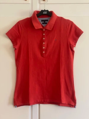 Röd pikétopp från Tommy Hilfiger - Klassisk röd pikétopp från Tommy Hilfiger med korta ärmar, krage och knappar framtill. Liten logga på bröstet och mjukt bomullsmaterial som känns skönt mot huden. Perfekt till jeans eller kjol för en sportig och stilren look.