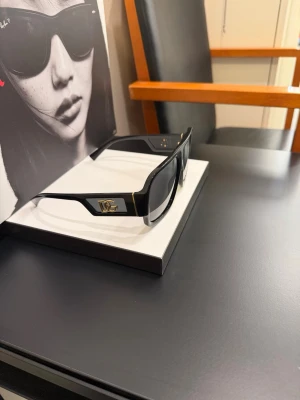 Svarta solglasögon från Dolce & Gabbana - Snygga svarta solglasögon från Dolce & Gabbana med breda bågar och guldiga DG-detaljer på sidorna. Rektangulär form och mörka glas som ger en cool och trendig look. Perfekta för dig som vill sticka ut med en exklusiv accessoar.