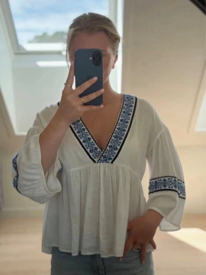 Vit boho-blus med blå brodyr - Säljer en luftig vit blus med blå broderade detaljer längs v-ringningen och ärmarna. Blusen har en avslappnad passform, vida trekvartsärmar och ett lätt, mjukt tyg som ger en bohemisk känsla. Perfekt till jeans eller shorts för en chill look.