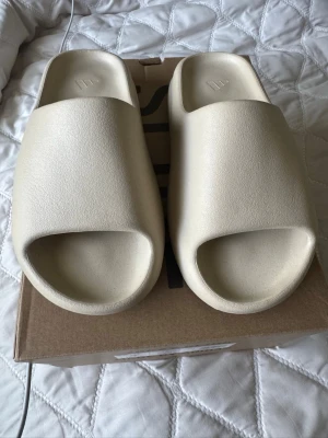 Beige Yeezy Slide tofflor (färgen bone) - Säljer ett par Yeezy Slide från Adidas i beige färg. Tofflorna har en chunky, modern design med öppen tå och räfflad sula för extra grepp. Tillverkade i ett mjukt och flexibelt skummaterial som gör dem riktigt bekväma att glida runt i.