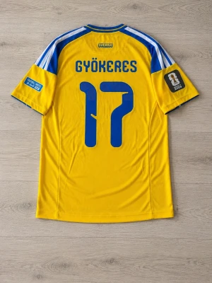 Sverige World cup jersey  - Gul och blå Sverige matchtröja från Adidas med Gyökeres och nummer 17 på ryggen. Tröjan har blå detaljer, officiella landslagsmärket på bröstet och CLIMACOOL-material för bra ventilation. Korta ärmar med tryck på båda och klassiska Adidas-ränder på axlarna.