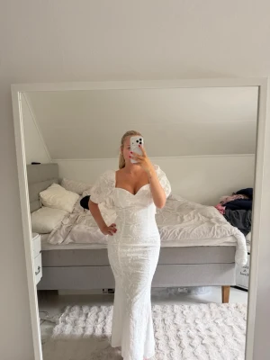 Vit långklänning med puffärmar - Elegant vit långklänning med puffiga kortärmar och v-ringad urringning från addore. Klänningen har figurnära passform och slits baktill för extra rörelsefrihet. Materialet har en lätt glittrig yta som ger en lyxig känsla. Perfekt för speciella tillfällen. Köpt för 1800kr i butik. Aldrig använt! 