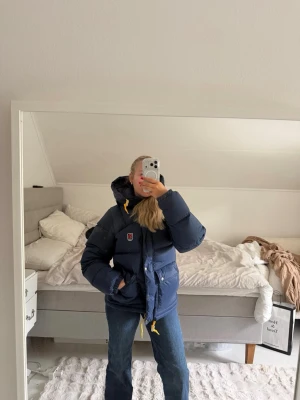 Mörkblå dunjacka från Fjällräven - Snygg mörkblå dunjacka från Fjällräven med huva och gula detaljer på snören och dragkedja. Jackan har två stora fickor framtill, justerbar midja och en klassisk Fjällräven-logga på bröstet. Perfekt för kalla vinterdagar och riktigt varm. Har endast använt någon gång. Skicket är som nytt. Köpte den för 7000kr i butik.