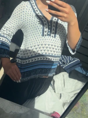Vit och blå blus med mönster - Säljer en luftig vit blus med blått mönster och dekorativa knappar framtill. Blusen har trekvartsärm och ett bohemiskt tryck med inslag av ränder och små blommor. Perfekt till jeans eller kjol för en avslappnad look.