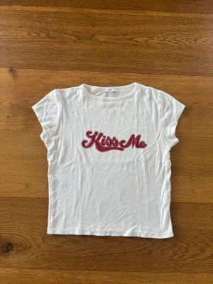 Vit t-shirt med röd text från Subdued - Säljer en vit t-shirt från Subdued med korta ärmar och rund hals. Framtill står det 'Kiss Me' i röd, glittrig text. Tillverkad i mjuk bomull, perfekt för en avslappnad och trendig look.