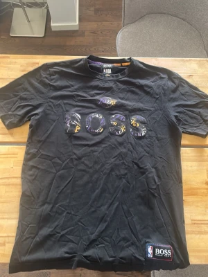 Svart Lakers x BOSS NBA t-shirt - Svart t-shirt från Hugo Boss i samarbete med NBA och Los Angeles Lakers. Stor Lakers-logga på bröstet och BOSS-tryck med lila och gula detaljer. Klassisk passform och korta ärmar. Tillverkad i mjuk bomull, perfekt för dig som gillar basket och streetwear.