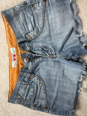 Blå lowiast jeansshorts från Rising - Säljer ett par lowais blå jeansshorts från Rising. En orange  insida som ger en cool detalj. Perfekta för varma dagar och enkel att matcha med allt.