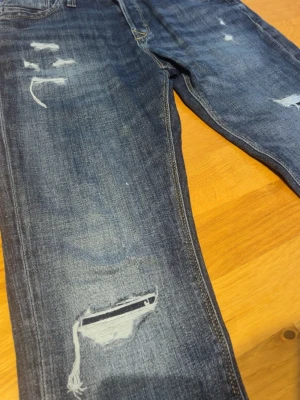 Jack&Jones jeans  - Säljer ett par blå jeans med slitningar och hål på benen för en trendig look. Jeansen har klassisk femficksmodell, normal midja och är tillverkade i denim med stretch. Perfekta för dig som gillar en avslappnad och cool stil.