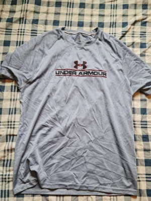 Grå Under Armour t-shirt - Snygg grå t-shirt från Under Armour med logga och text på bröstet. Tillverkad i ett mjukt och lätt material som andas, perfekt för träning eller chill. Klassisk passform med korta ärmar och rund halsringning.