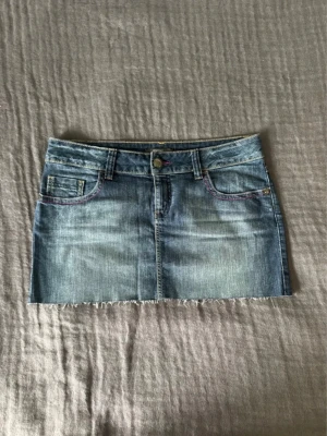 Blå jeanskjol med rå kant - Säljer en klassisk blå jeanskjol i kort modell med rå, fransig kant nertill. Kjolen har fem fickor, bälteshällor och knappstängning framtill. Snygga kontrastsömmar och slitningar ger en cool vintagekänsla. Perfekt till sneakers eller boots!