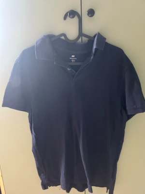 Mörkblå pikétröja från H&M - Klassisk mörkblå pikétröja från H&M i regular fit. Tröjan har korta ärmar, krage och knappar vid halsen. Tillverkad i mjuk bomull som känns skön mot huden. Perfekt för en avslappnad och stilren look.