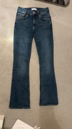 Blå bootcut jeans  - Säljer ett par klassiska blå bootcut jeans från Gina. Jeansen har fem fickor, normalhög midja och snygga slitningar framtill. Tillverkade i stretchig denim för skön passform och med kopparfärgade detaljer vid fickorna. De är uppsydda för att passa under 164cm