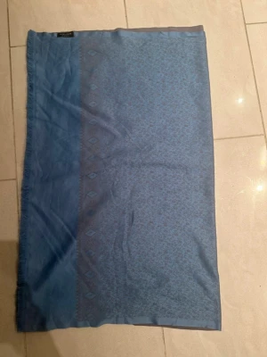 Blå mönstrad halsduk  - Snygg blå halsduk  med diskret mönster och fransad kant. Halsduken är rektangulär och har en elegant bård längs ena sidan. Tillverkad i ett tunt, mjukt material som känns skönt mot huden. Perfekt för att lyfta din outfit.