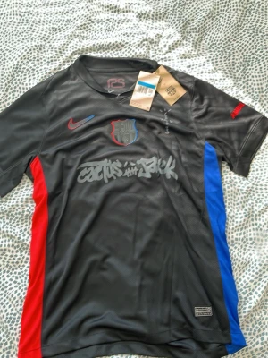 Svart FC Barcelona Nike Dri-Fit tröja - Snygg svart FC Barcelona tröja från Nike med Dri-Fit material. Tröjan har klubbmärke på bröstet, röda och blå detaljer på sidorna samt tryck på bröstet. Perfekt för träning eller att bära till vardags. Officiell produkt med etikett kvar.