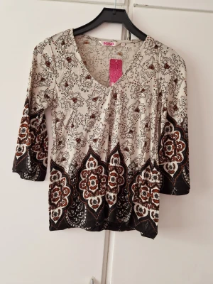 Mönstrad blus från Indiska - Säljer en snygg blus från Indiska med blommigt mönster i beige, svart och brunt. Blusen har v-ringning och trekvartsärm, perfekt för dig som gillar boho-vibbar och detaljerade prints. Superskön och lätt att styla till jeans eller kjol.