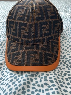 Fendi keps med FF-mönster - Säljer en snygg keps från Fendi med det klassiska FF-logomönstret i svart och brunt. Skärmen har en brun läderdetalj som ger en exklusiv känsla. Justerbar baktill och fodrad med svart tyg på insidan. Perfekt för dig som gillar lyxiga accessoarer.