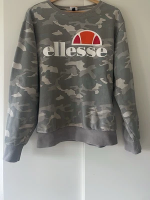 Grå camo sweatshirt från Ellesse - Snygg grå sweatshirt från Ellesse med camouflagemönster och stor logga framtill i vitt, orange och rött. Tröjan har rund halsringning, ribbade muddar vid ärmslut och nederkant. Perfekt för en avslappnad streetwear-look. Något mindre i storleken så är mer som en  L i storleken. 