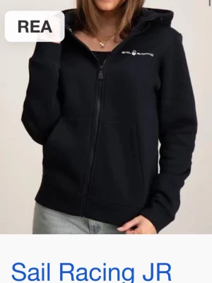 Svart hoodie från Sail Racing - Snygg svart hoodie från Sail Racing med dragkedja framtill och huva. Diskret logga på bröstet och stora fickor framtill. Perfekt för en avslappnad och sportig stil. Tillverkad i mjukt material som känns skönt mot huden.