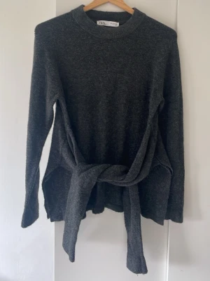 Mörkgrå stickad tröja från Zara - Mysig mörkgrå stickad tröja från Zara med långa ärmar och rund halsringning. Tröjan har en unik knytning framtill som ger en cool detalj. Perfekt till höst och vinter för en avslappnad men trendig look.