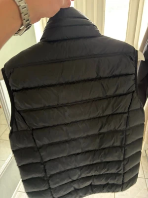 Svart dunväst från Emporio Armani EA7 - Snygg svart dunväst från Emporio Armani EA7 med quiltad design och hög krage. Den är använd fåtal gånger Västen har dragkedja framtill och två sidofickor. EA7-logga i vitt på bröstet ger en clean och sportig look. Perfekt för lager-på-lager och streetstyle. OBS hämtas på plats, skickas ej