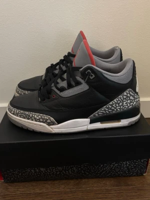 Air Jordan 3 Retro OG Black Cement - Säljer ett par Air Jordan 3 Retro OG 'Black Cement' med svart läder, grå mesh och ikoniska elefantmönster på tå och häl. Skorna har röda detaljer på plösen och klassisk Nike Air-logga bak. Perfekta för dig som gillar streetwear och sneakerskultur.