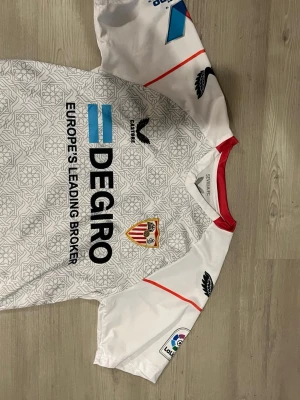 Almería vit matchtröja Hummel - Säljer en vit matchtröja från Almería med röda detaljer och klubbmärke på bröstet. Tröjan har tryck från DEGIRO och La Liga-logga på ärmen. Tillverkad i lätt och ventilerande material, perfekt för fotboll eller träning.