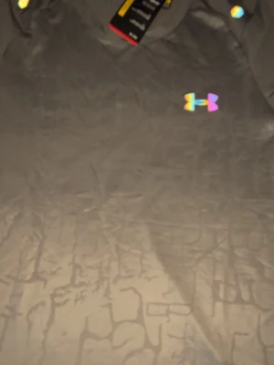 Grå mönstrad t-shirt från Under Armour - Snygg grå t-shirt från Under Armour med diskret mönster i tyget och ett regnbågsfärgat logotryck på bröstet. Tillverkad i ett lätt och funktionellt syntetmaterial som passar perfekt till träning. Klassisk passform och korta ärmar.