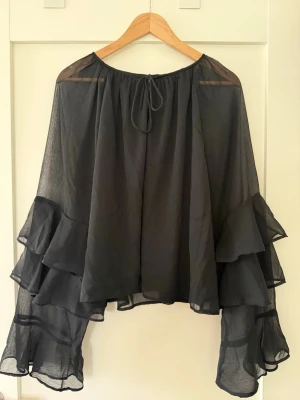 Svart volangblus med vida ärmar Nelly  - Säljer en svart transparent blus med vida, långa ärmar och flera lager volanger. Blusen har rund halsringning med knytband och är tillverkad i ett tunt, flowy material som ger en riktigt snygg och dramatisk look.