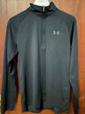 Svart träningströja från Under Armour - Svart långärmad träningströja från Under Armour med halv dragkedja framtill och hög krage. Tillverkad i ett stretchigt funktionsmaterial som andas. Diskret logga på bröstet och i nacken. Perfekt för löpning eller gym.