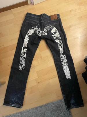 Evisu jeans - Säljer ett par mörkblå Evisu jeans med unikt vitt grafiskt tryck på bakfickorna och längs benen. Klassisk femficksmodell med raka ben och knappgylf. Jeansen har en cool streetwear-vibe och är tillverkade i kraftigt denimtyg.