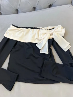 Svart offshoulder topp med rosett - Trendig svart offshoulder topp med lång ärm och bred, krämvit volangkant upptill. Stor dekorativ rosett i samma krämvita tyg ger en extra stylish touch. Perfekt för dig som vill sticka ut med en unik detalj på din outfit.