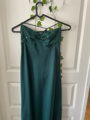 Grön långklänning med paljetter - Mörkgrön långklänning med smala axelband och paljettprydd överdel. Klänningen har en söt rosettdetalj framtill och är gjord i ett glansigt material som ger en elegant look. Perfekt för bal eller liknande 😁