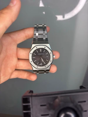 Audemars Piguet Royal Oak klocka - Snygg Audemars Piguet Royal Oak klocka med mörkgrå urtavla och datumvisning. Klockan har en ikonisk åttakantig boett och länkarmband i silverfärgat stål. Perfekt för dig som vill ha en stilren och lyxig accessoar.