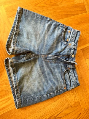 Blå denimshorts med hög midja - Klassiska blå jeansshorts i denim med hög midja och fem fickor. Shorts med dragkedja och knapp framtill, samt bälteshällor. Perfekta för sommaren och ger en avslappnad vibe till din outfit.