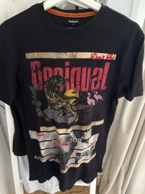 Desigual Shirt - Cool svart t-shirt från Desigual med stort färgglatt tryck framtill som visar en tiger, flamingos och grafiska detaljer. T-shirten har rund hals, korta ärmar och är gjord i mjuk bomull. Perfekt för dig som gillar unika prints och streetstyle.