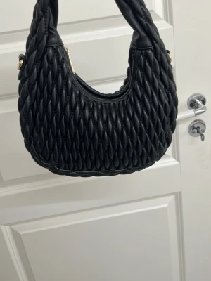 Svart handväska från Don Donna - Snygg svart handväska från Don Donna med flätad design och rundad form. Väskan är gjord i konstläder och har guldfärgade detaljer vid handtaget. Perfekt accessoar för att lyfta din outfit.