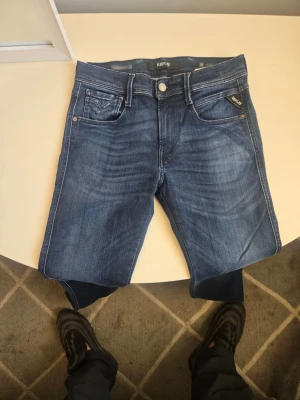 Blå jeans från Replay W28 L32 - Snygga blå jeans från Replay med klassisk femficksdesign och diskreta slitningar. Jeansen har raka ben och tydliga kontrastsömmar. Tillverkade i mjukt denimtyg som ger en bekväm passform. Perfekta för en avslappnad och trendig look.