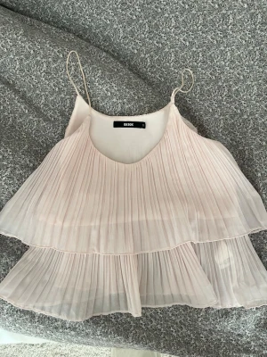 Ljusrosa plisserad topp från BikBok - Säljer en ljusrosa plisserad topp från BikBok med tunna axelband och dubbla volanger. Toppen har ett luftigt och flowy material som ger en romantisk vibe. Perfekt för sommardagar eller när du vill ha en söt och enkel look.