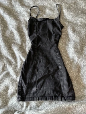 Svart jeansklänning med smala axelband - Snygg svart kort jeansklänning med smala axelband och figurnära passform. Klänningen har sömdetaljer framtill och är perfekt för en trendig och avslappnad look. Passar dig som gillar enkel men stilren design.