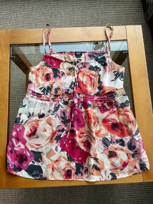 Blommigt linne från Esprit - Supersött linne från Esprit med blommigt mönster i rosa, orange och svart.Tunt, luftigt tyg med smala reglerbara axelband. Knyts framtill.. Storlek 32/34. 