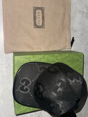 Svart keps från Gucci med GG-mönster - Snygg svart keps från Gucci med det klassiska GG-monogrammet över hela kepsen. Tillverkad i mesh-liknande material med justerbar rem bak. Kepsen kommer med original dustbag och grön Gucci-box. Perfekt för dig som gillar exklusiva accessoarer.