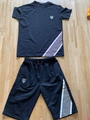 Svart set med vita ränder - Säljer ett svart tvådelat set med t-shirt och shorts. Båda delarna har vita diagonala ränder och en liten sköldlogga på bröstet och benet. Setet har en sportig look och är gjort i mjukt bomullsliknande material med resår och dragsko i midjan på shortsen.