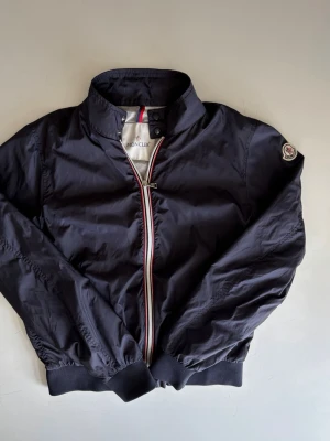 Moncler Bomber Jacket - Snygg svart vindjacka från Moncler med hög krage och ribbade muddar. Jackan har dragkedja med röd, vit och blå detalj samt Moncler-logga på ärmen. Storlek 13-15 år. Nypris : ca 4000 SEK Pris : 1500 SEK