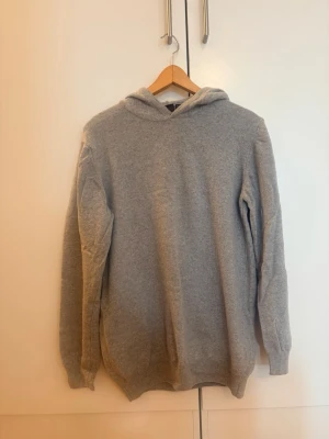 Grå hoodie från Oscar Jacobson - Mjuk och stilren grå hoodie från Oscar Jacobson, tillverkad i Italien. Hoodien har en klassisk huva och är gjord i ett skönt material som passar perfekt för en avslappnad look. Enkel design utan tryck eller dragkedja, med ribbade muddar vid ärmslut och nederkant.