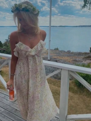 Blommig långklänning med volang - Romantisk långklänning med smala axelband och volangdetalj vid bysten. Klänningen har ett ljust blommigt mönster i pastellfärger och är perfekt för somriga tillfällen. Tillverkad i ett lätt och luftigt material som ger en skön känsla.