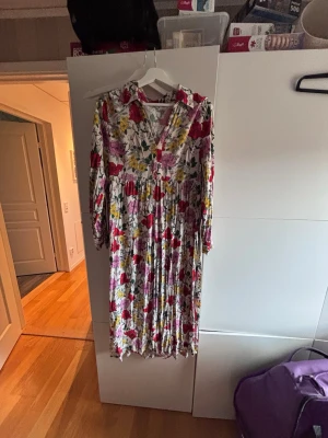 Blommig långklänning med krage - Säljer en långklänning med färgglatt blommönster i rött, gult och lila på vit botten. Klänningen har v-ringning, krage och långa ärmar. Perfekt för dig som gillar en romantisk och bohemisk stil.