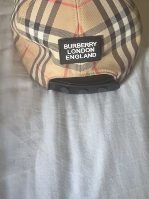 Burberry keps  - Snygg beige keps från Burberry med klassiskt rutmönster i svart, vitt och rött. Kepsen har justerbar snapback och en tydlig Burberry London England-patch bak. Tillverkad i bomull och polyester för en skön och stilren look.